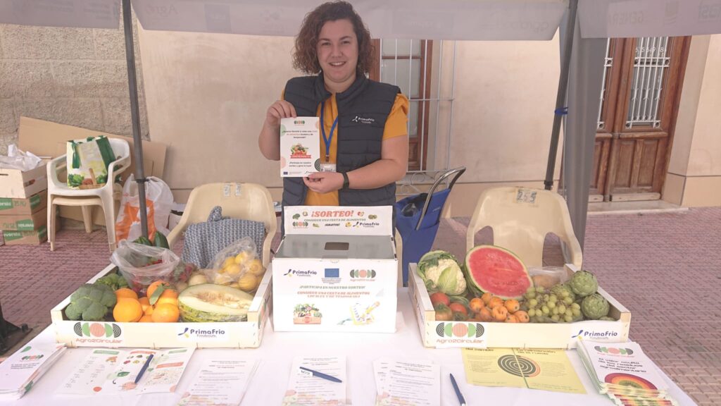 Una chica en un puesto informativo, entre dos cajas de frutas y verduras, ofrece un folleto a quien hace la fotografía